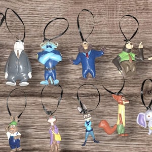May include: A collection of Zootopia character ornaments, each with a black loop for hanging. The ornaments feature characters like Judy Hopps, Nick Wilde, and Flash, in various poses and outfits. The ornaments are made of plastic and are colorful.