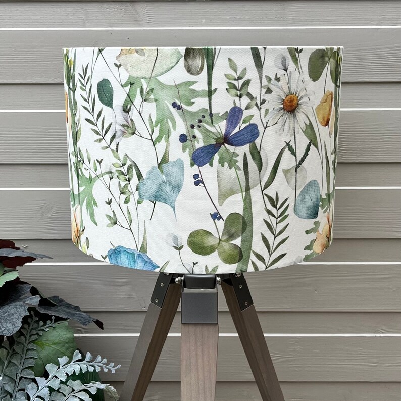 A white lampshade with a floral pattern. The pattern features green leaves, blue flowers, and yellow flowers. The lampshade is on a tripod stand with dark brown legs.
