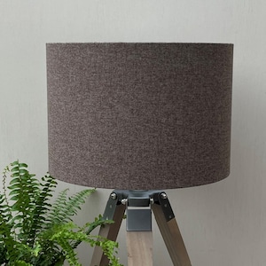 May include: A cylindrical lampshade in a textured dark brown fabric sits atop a tripod base. The base features three light-toned wooden legs connected by dark grey metal hardware, providing a stable structure for the overhead lighting fixture.