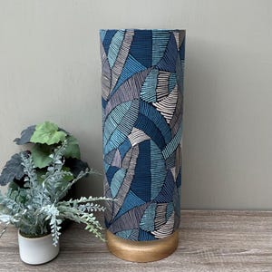 May include: A cylindrical table lamp featuring a fabric shade with a repeating abstract pattern of blue, teal, and grey curved lines. The lamp has a circular light-coloured wooden base and stands upright, providing a structured and geometric appearance.