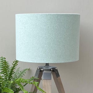 May include: A table lamp featuring a cylindrical lampshade in a soft, textured mint green fabric. The base is constructed from three wooden legs connected by a dark grey metal bracket, providing a stable tripod structure for the light fixture.