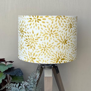 May include: A cylindrical lamp shade featuring a repeating pattern of mustard yellow, brushstroke-style floral bursts on a white background. The shade is mounted on a tripod base with three wooden legs and silver-toned metal hardware connectors.