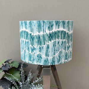 May include: A cylindrical lampshade featuring a repeating vertical pattern of watercolour-style brushstrokes in varying shades of teal, turquoise, and light blue against a crisp white background, mounted on a wooden tripod base.