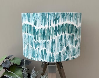 May include: A cylindrical lamp shade featuring a repeating vertical pattern of watercolor-style brushstrokes in varying shades of teal, turquoise, and light blue against a crisp white background, mounted on a wooden tripod base.