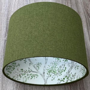 May include: A cylindrical drum lampshade featuring a solid olive green fabric exterior with a textured weave. The interior lining displays a white background printed with a repeating pattern of light grey tree branches and small green leaves.