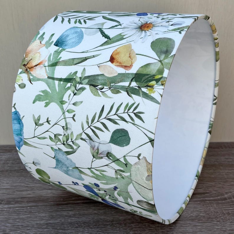 A white lampshade with a floral pattern. The pattern features green leaves, blue, yellow, and white flowers. The lampshade is drum shape and has a white interior.