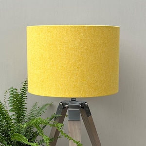 May include: A bright yellow lampshade, with a textured finish, is mounted on a wooden tripod lamp base. The lamp is positioned next to a green fern. The lampshade is a cylindrical shape. The lamp is a home decor item, suitable for a living room.