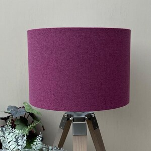 May include: A cylindrical lampshade in a deep magenta purple fabric with a subtle heathered texture. The shade is mounted on a tripod base featuring three light-toned wooden legs connected by a central silver-coloured metal bracket assembly.