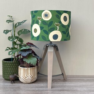 May include: A table lamp with a cylindrical green fabric shade featuring a cream and dark blue floral pattern. The lamp has a light-coloured wooden tripod base with metal hardware. The lamp is positioned next to two potted plants and a small bird figure.