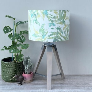 May include: A table lamp with a cylindrical shade featuring a green, yellow, and teal botanical leaf pattern. The lamp has a tripod base made of light-coloured wood with silver metal hardware connecting the three legs to the central lamp socket.