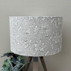 May include: A grey lampshade featuring a white thistle design. The lampshade is cylindrical and rests on a wooden tripod stand. The thistle pattern is detailed and covers the entire shade, creating a botanical theme.