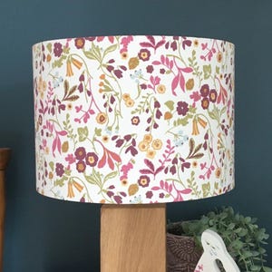 May include: A cylindrical lampshade featuring a white background with a repeating botanical pattern of small flowers and leaves in shades of magenta, mustard yellow, sage green, and light blue, mounted on a light-toned wooden lamp base.