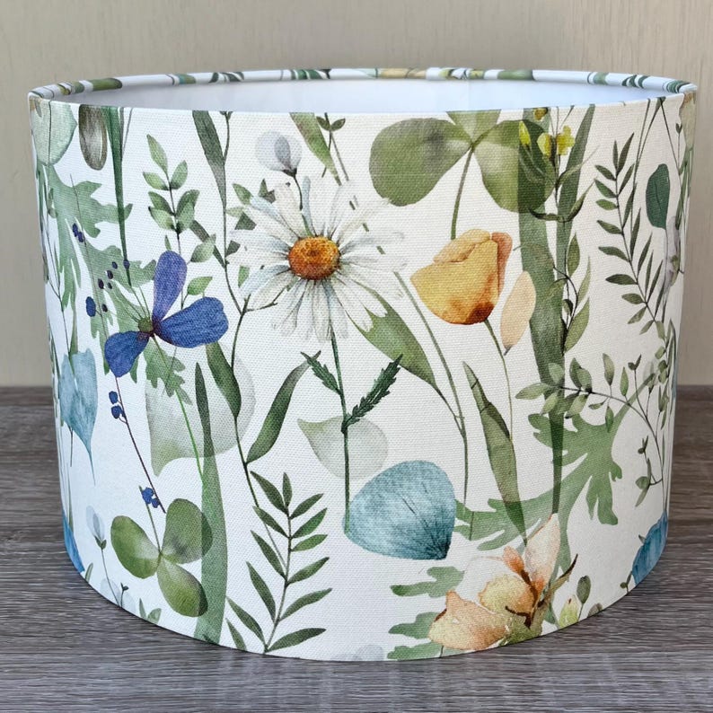 A white drum lampshade with a floral pattern. The pattern features green leaves, blue flowers, and yellow flowers. The lampshade is made of fabric and has a white background.