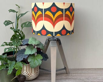 May include: A table lamp with a cylindrical shade featuring a repeating pattern of cream, dark blue, orange, yellow, and brown stylized floral shapes. The lamp has a light-colored wooden tripod base with silver metal hardware and three legs.