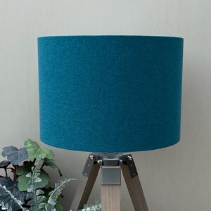 May include: A cylindrical lampshade in a solid teal blue fabric with a visible woven texture. The shade is mounted on a tripod base featuring three light-toned wooden legs connected by a central metal bracket with silver-coloured hardware.