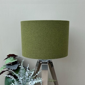 May include: A cylindrical lamp shade in a solid olive green fabric with a textured weave. The shade is mounted on a tripod base featuring three light-toned wooden legs connected by silver-coloured metal hardware at the top junction point.