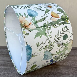 A white drum lampshade with a floral pattern. The pattern features green leaves, blue flowers, and yellow flowers. The lampshade is made of fabric and has a white lining.