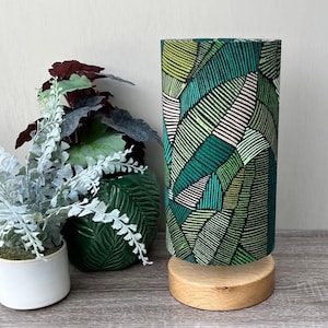 May include: A cylindrical table lamp featuring a fabric shade with a geometric pattern of green, lime, and white stripes. The lamp is supported by a circular, light-coloured wooden base. The design consists of angular, leaf-like shapes.