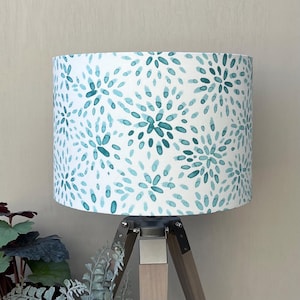 May include: A cylindrical lamp shade featuring a white background with a repeating pattern of teal and light blue brushstroke petals arranged in circular floral clusters. The shade is mounted on a tripod base made of dark wood with metal accents.