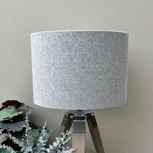 May include: A cylindrical lamp shade with a heathered light grey fabric texture. The shade is mounted on a tripod base featuring three wooden legs connected by a dark grey metal hub at the top, providing a stable structure for the light fixture.