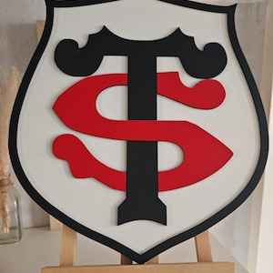 Peut inclure: Un écusson en bois en forme de bouclier avec un fond blanc, une bordure noire et un logo rouge et noir. Le logo présente un design stylisé "T" et "S". L'écusson est exposé sur un petit chevalet en bois.