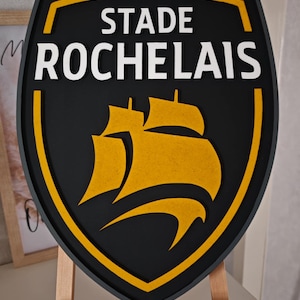 Peut inclure: Un panneau noir et jaune en forme de bouclier avec les mots "STADE ROCHELAIS" en blanc. Le panneau présente un emblème jaune de deux voiliers. Le panneau est exposé sur un chevalet en bois.