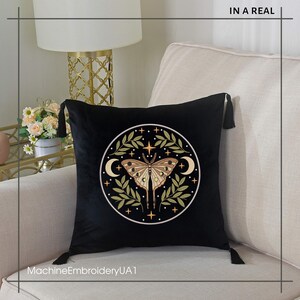 May include: Black velvet throw pillow with a circular embroidered design featuring a moth, crescent moons, stars, and olive-green leaves. The pillow has black tassels on each corner. The text "IN A REAL" is at the top and "MachineEmbroideryUA1" at the bottom.