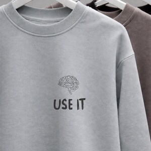 Może przedstawiać: Jasnoszara bluza z czarną grafiką m&oacute;zgu i napisem "USE IT" poniżej. Bluza wisi na białym wieszaku. W tle widoczne są inne bluzy w kolorze brązowym i czarnym.