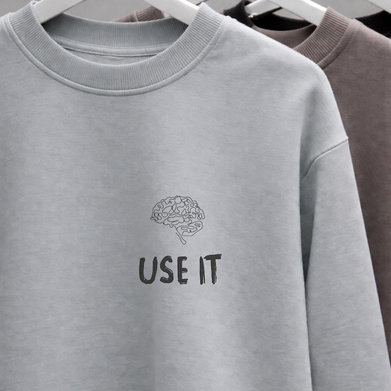 Może przedstawiać: Jasnoszara bluza z czarną grafiką m&oacute;zgu i napisem "USE IT" poniżej. Bluza wisi na białym wieszaku. W tle widoczne są inne bluzy w kolorze brązowym i czarnym.