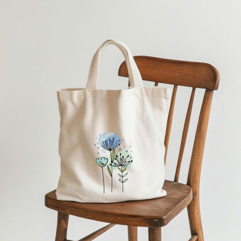 May include: Cream-colored tote bag with a floral design. The design features blue, green, and teal watercolor flowers with black outlines. The bag has two handles and is sitting on a wooden chair.