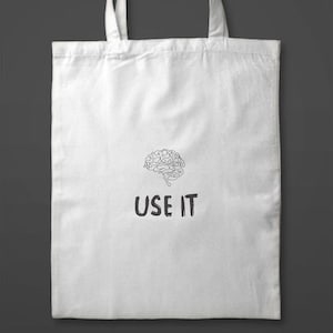 Może przedstawiać: Biała torba typu tote z czarną grafiką m&oacute;zgu i napisem "USE IT" poniżej. Torba ma dwa białe uchwyty i jest wykonana z miękkiego, trwałego materiału. Projekt jest prosty i przyciągający wzrok.