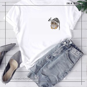 Op de afbeelding: Wit T-shirt met een schattige cartoon-uil die een gestreepte slaapmuts draagt. De uil is geborduurd op het shirt. Het shirt ligt op een houten oppervlak met een paar grijze schoenen en een blauwe denim short.