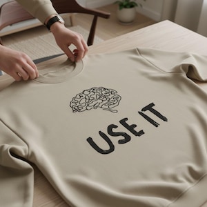 Może przedstawiać: Beżowa bluza z czarną grafiką m&oacute;zgu i napisem "USE IT" poniżej. Bluza wykonana jest z miękkiego materiału i ma okrągły dekolt. Zdjęcie przedstawia bluzę na drewnianej powierzchni.