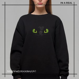 Pu&ograve; includere: Felpa nera con un designo ricamato di occhio di drago verde. Il design &egrave; centrato sulla parte anteriore della felpa.
