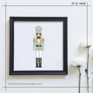 May include: Framed embroidery art of a nutcracker soldier in a black frame. The nutcracker is light blue with gold and white accents, black boots, and a detailed mustache. The text "in a real" is in the top right corner. The text "MachineEmbroideryUA1" is at the bottom.