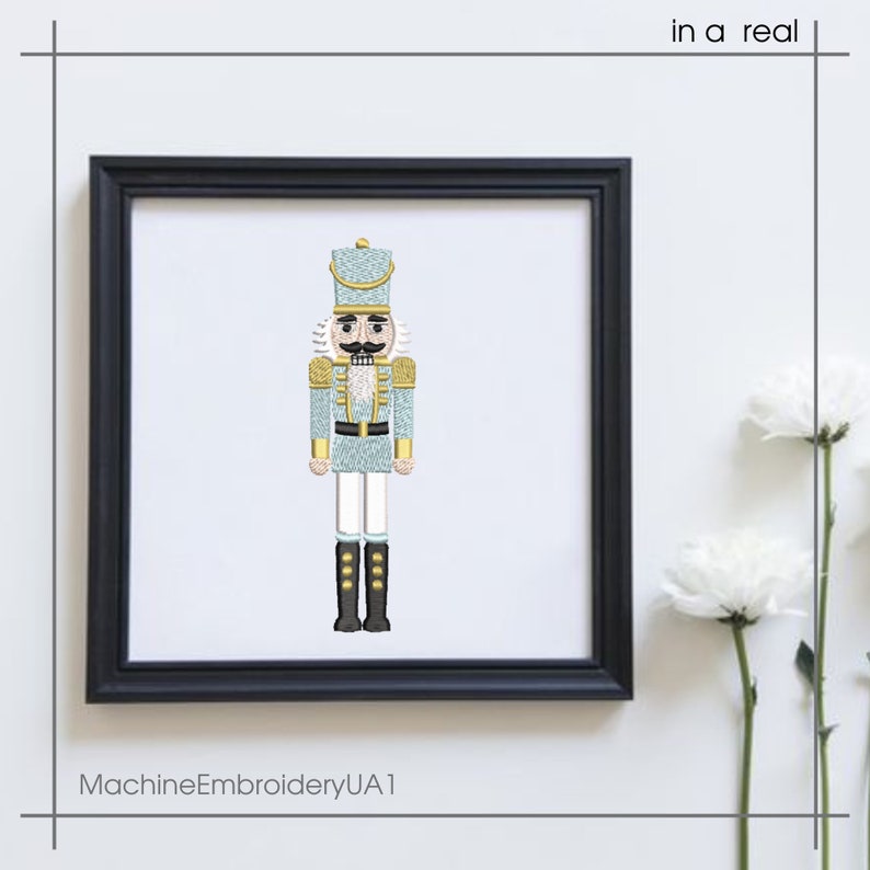 May include: Framed embroidery art of a nutcracker soldier in a black frame. The nutcracker is light blue with gold and white accents, black boots, and a detailed mustache. The text "in a real" is in the top right corner. The text "MachineEmbroideryUA1" is at the bottom.