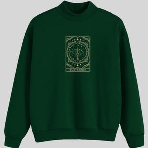 Pu&ograve; includere: Una felpa verde scuro con una grafica dorata di un simbolo zodiacale, una freccia e un arco, e la parola "Sagittario".