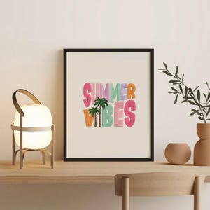 May include: A framed print with the words "SUMMER VIBES" in colorful, textured letters. The artwork includes palm trees and is set against a cream background. The frame is black, and the print is displayed on a wooden surface.