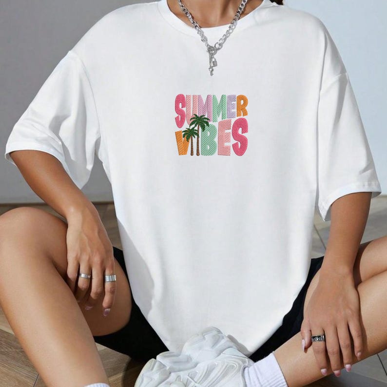 May include: White t-shirt with the text "SUMMER VIBES" in colorful, embroidered letters and a palm tree graphic. The shirt has short sleeves and a relaxed fit, perfect for warm weather.