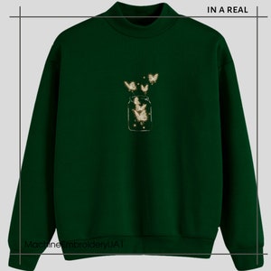 May include: A dark green sweatshirt with a white embroidered design of a jar with butterflies flying out of it. The text "IN A REAL" is in the top right corner of the image.
