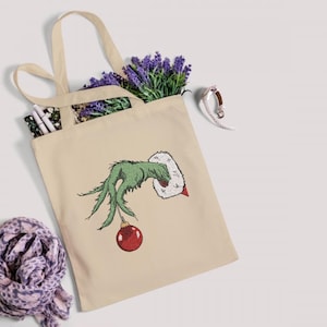 May include: A beige canvas tote bag with a green hand holding a red Christmas ornament. The hand is wearing a white furry cuff. The Grinch's hand is embroidered on the bag.