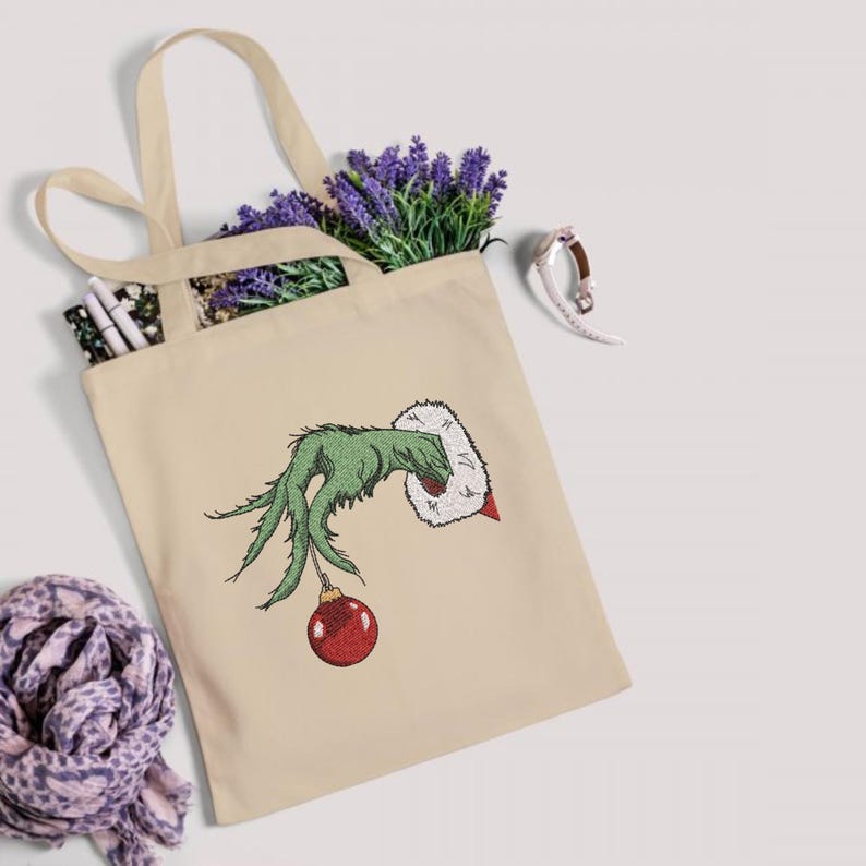 May include: A beige canvas tote bag with a green hand holding a red Christmas ornament. The hand is wearing a white furry cuff. The Grinch's hand is embroidered on the bag.