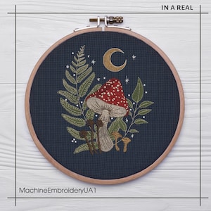 May include: A circular embroidery hoop with a dark blue background featuring a red and white mushroom with white spots, surrounded by green leaves and a crescent moon. The hoop is made of wood and has a natural finish.