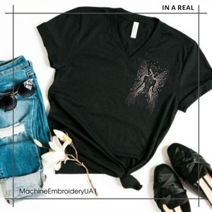May include: Black V-neck t-shirt with a white outline of a fairy with wings and stars. The text "IN A REAL" is in the top right corner of the image.