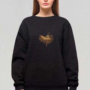 May include: Black sweatshirt featuring a gold-colored illustration of a ballerina in a flower-like design. The dancer is in a graceful pose, with arms and legs extended. The design is centered on the chest.