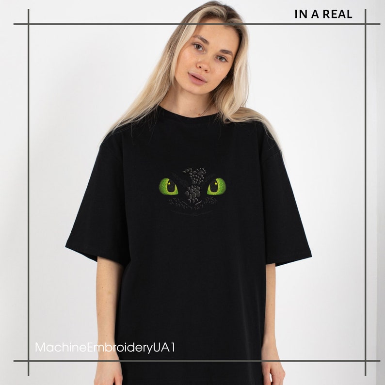 Pu&ograve; includere: Maglietta nera con una grafica di occhio di drago verde. Il testo "IN A REAL" &egrave; nell'angolo in alto a destra dell'immagine. Il testo "MachineEmbroideryUA1" &egrave; nell'angolo in basso a sinistra dell'immagine.