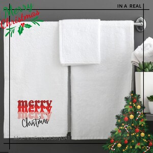 May include: Two white towels hanging on a rack. The larger towel features embroidered text that reads merry merry merry Christmas in stacked red, pink, and black script lettering. A small white hand towel is folded and draped over the rack.