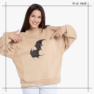 May include: A beige crewneck sweatshirt featuring a black embroidered graphic of a bat with spread wings on the center chest. The text "in a real" is visible in the top right corner, and "MachineEmbroideryUA1" is at the bottom left.