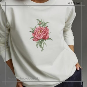 May include: White sweatshirt with a pink embroidered flower design. The text "IN A REAL" is printed on the top right corner of the sweatshirt. The text "MachineEmbroideryUA1" is printed on the bottom left corner of the sweatshirt.