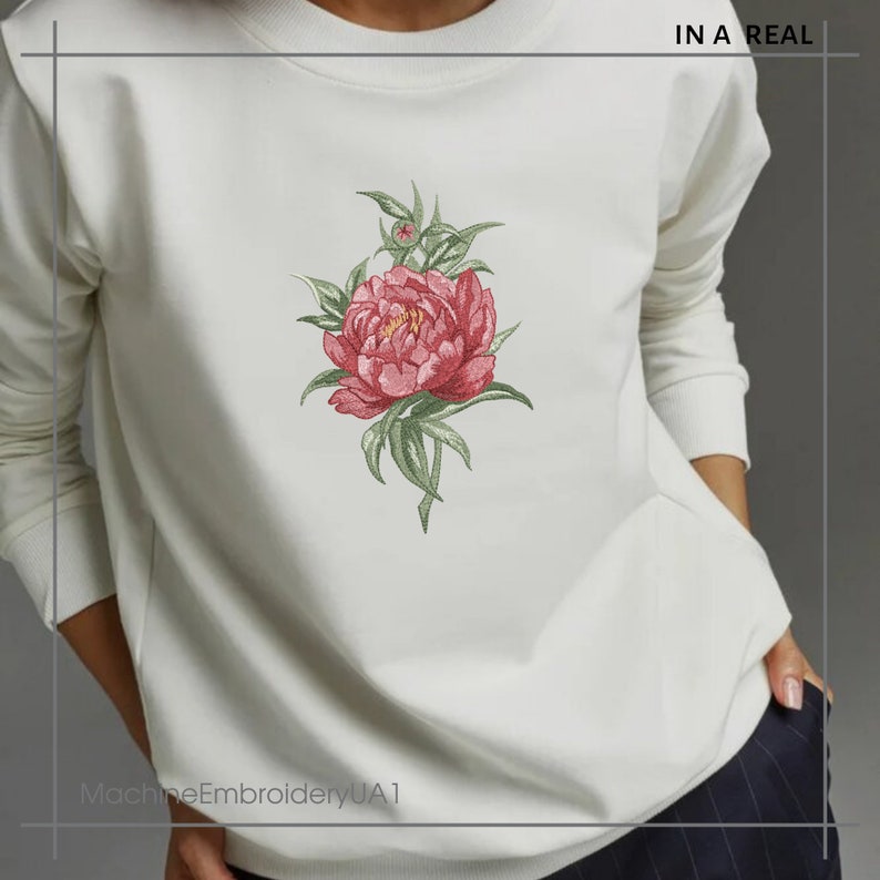 May include: White sweatshirt with a pink embroidered flower design. The text "IN A REAL" is printed on the top right corner of the sweatshirt. The text "MachineEmbroideryUA1" is printed on the bottom left corner of the sweatshirt.